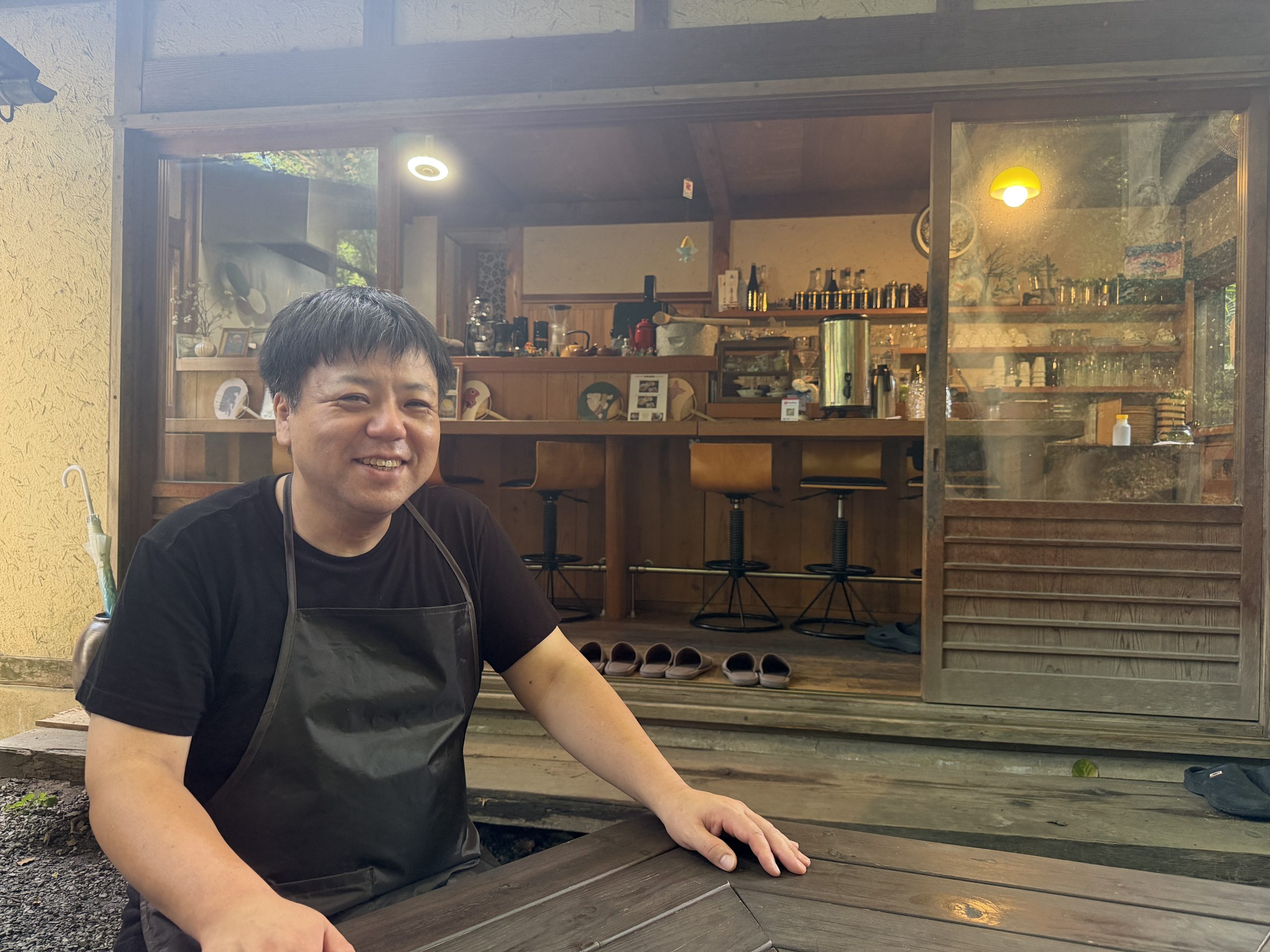 縁側のテーブル席で、店主らしき男性が笑顔で座っている様子。