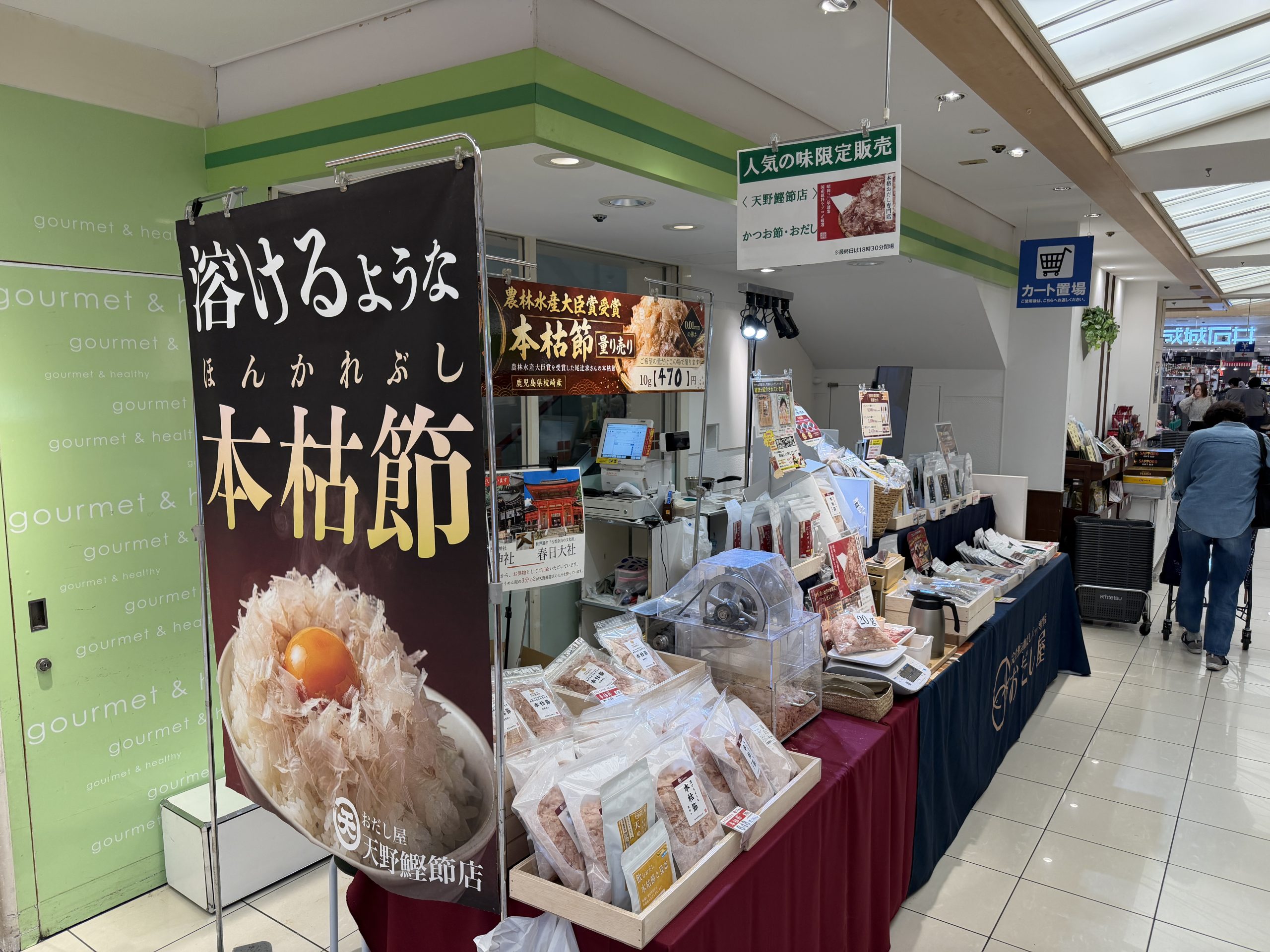 百貨店の食品売り場に出店している鰹節店の催事コーナー。大きな本枯節ののぼりと、鰹節やだし関連商品が並んだ売り場の様子。
