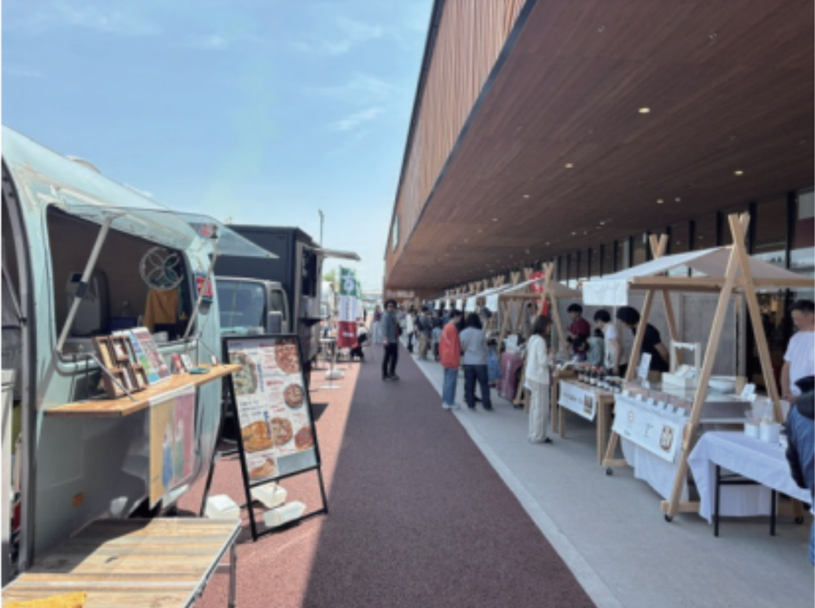 キッチンカーや木製屋台が並ぶイベント会場の通路で、買い物を楽しむ人たちでにぎわっている様子。