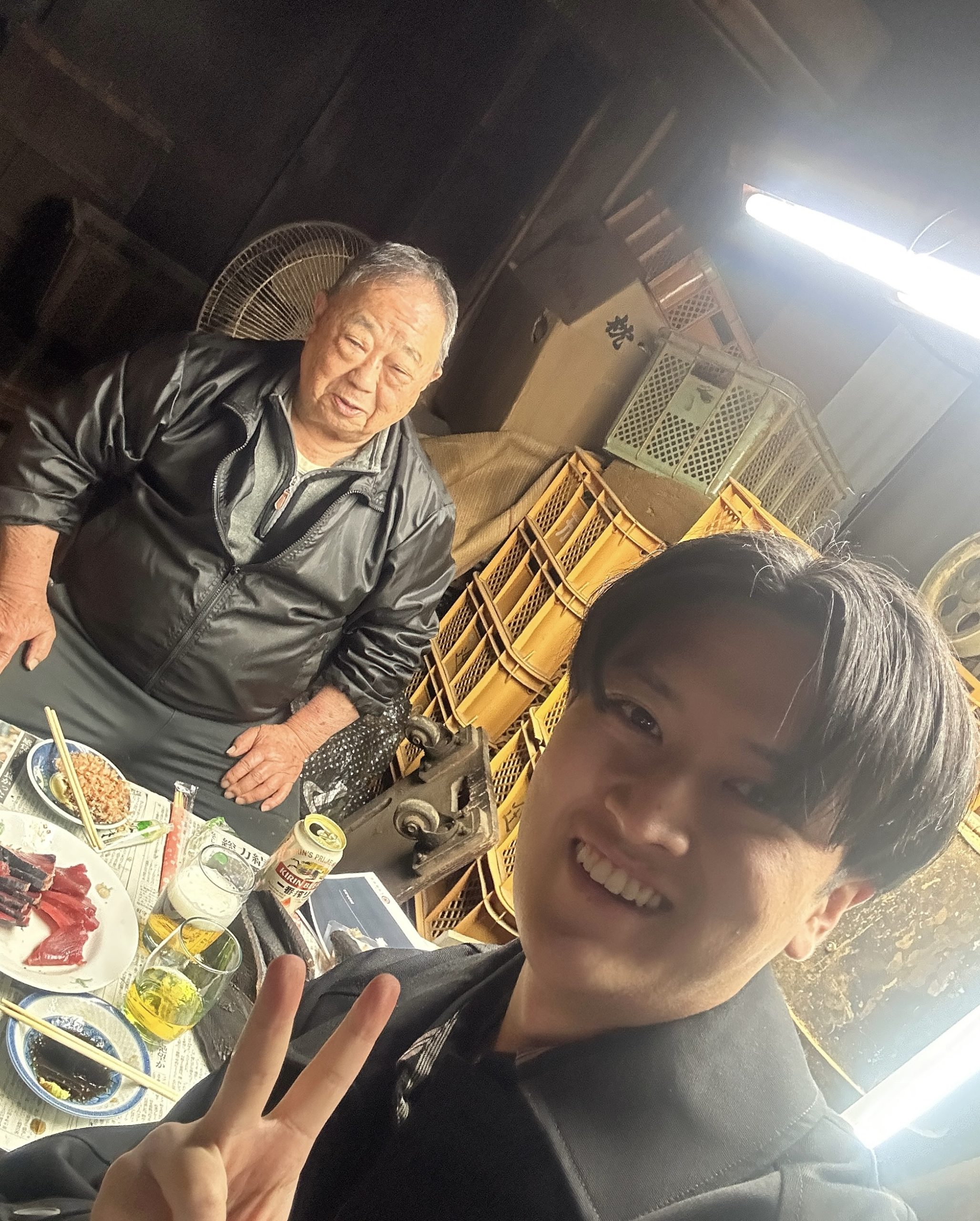 作業場で先代らしき男性と若い男性が、刺身や飲み物を囲んで談笑している様子